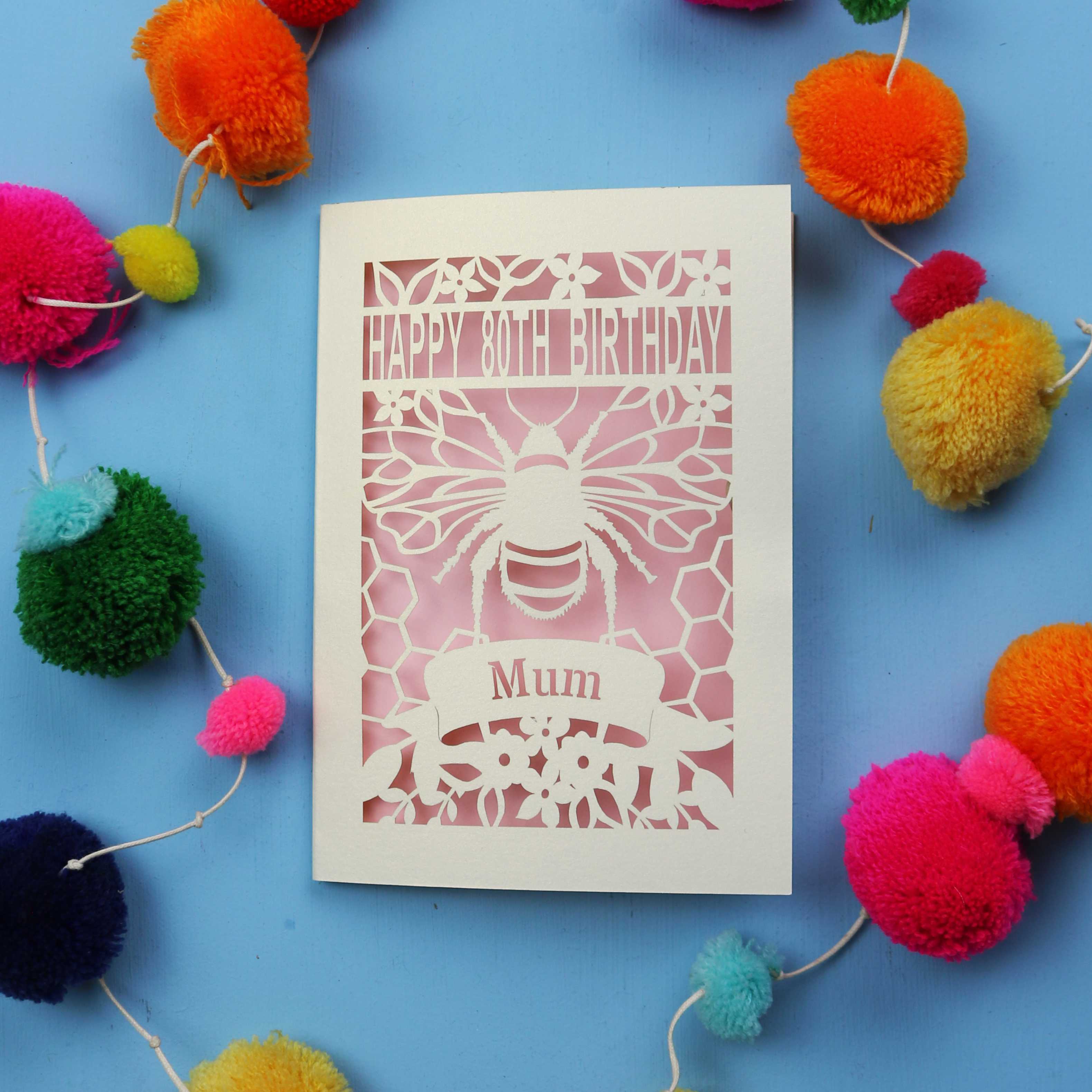 Laser-cut birthday card with bee design and 'Happy 80th Birthday Mum' text on a blue background with colourful pom-poms.