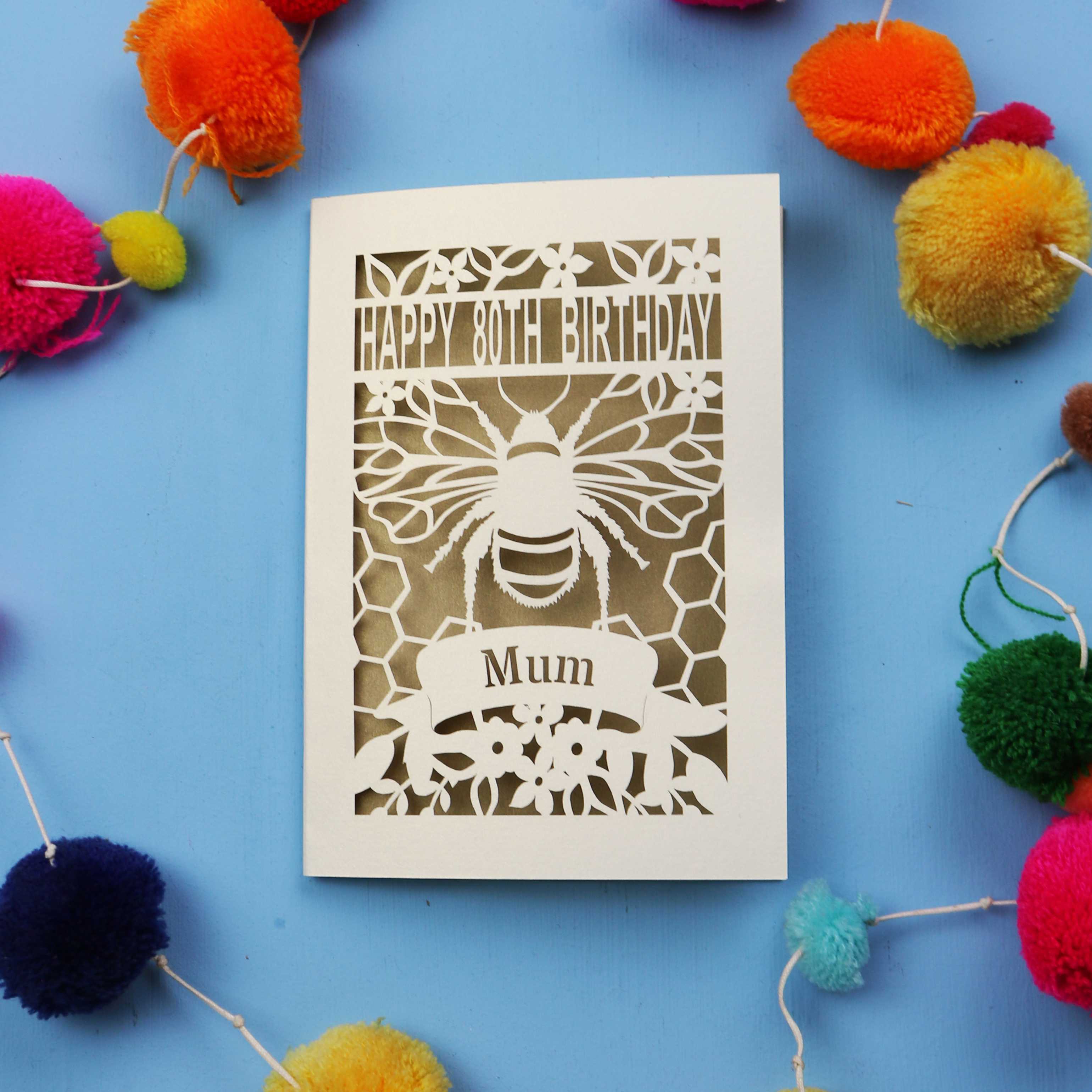 Laser-cut birthday card with bee design and 'Happy 80th Birthday Mum' text on a blue background with colourful pom-poms.