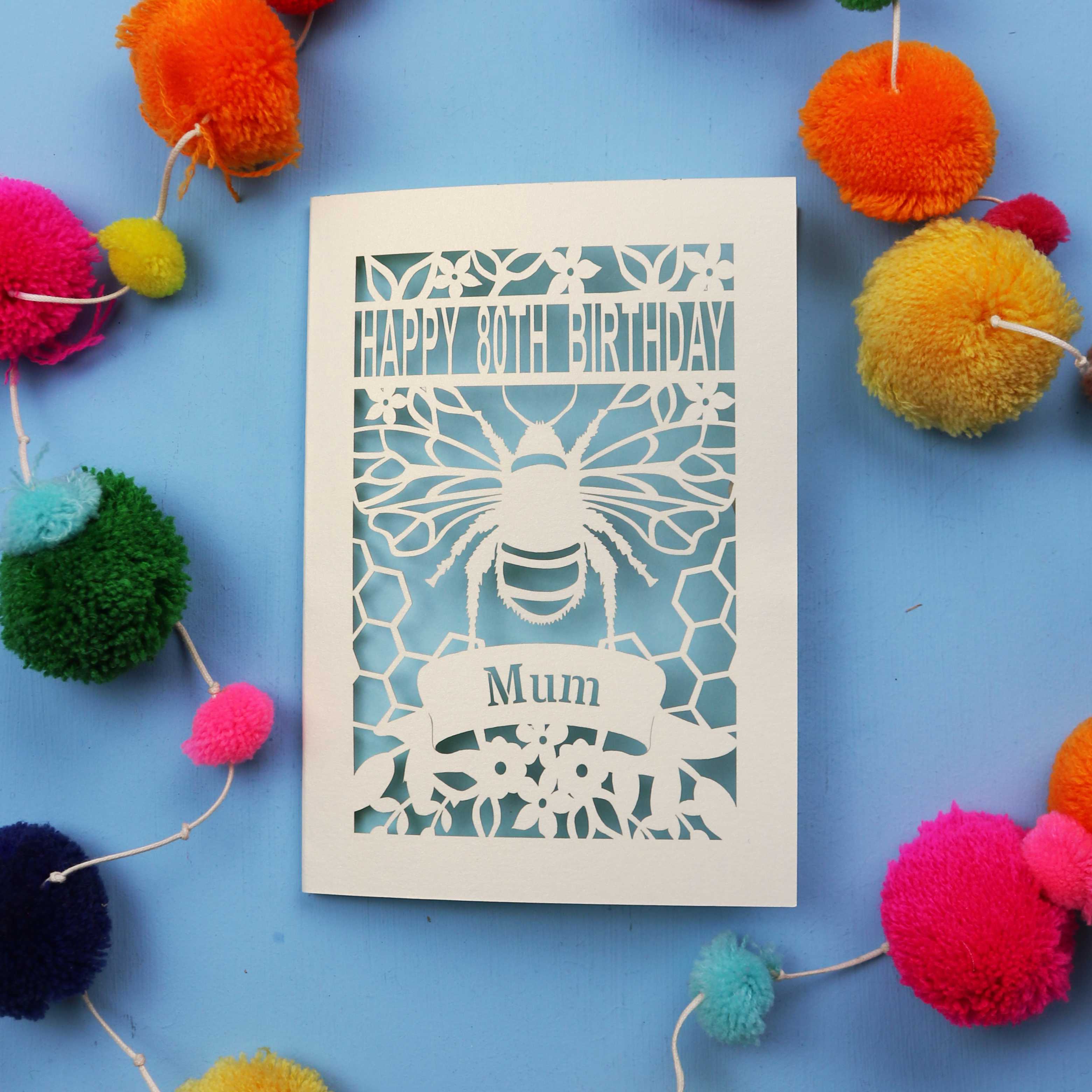 Laser-cut birthday card with bee design and 'Happy 80th Birthday Mum' text on a blue background with colourful pom-poms.