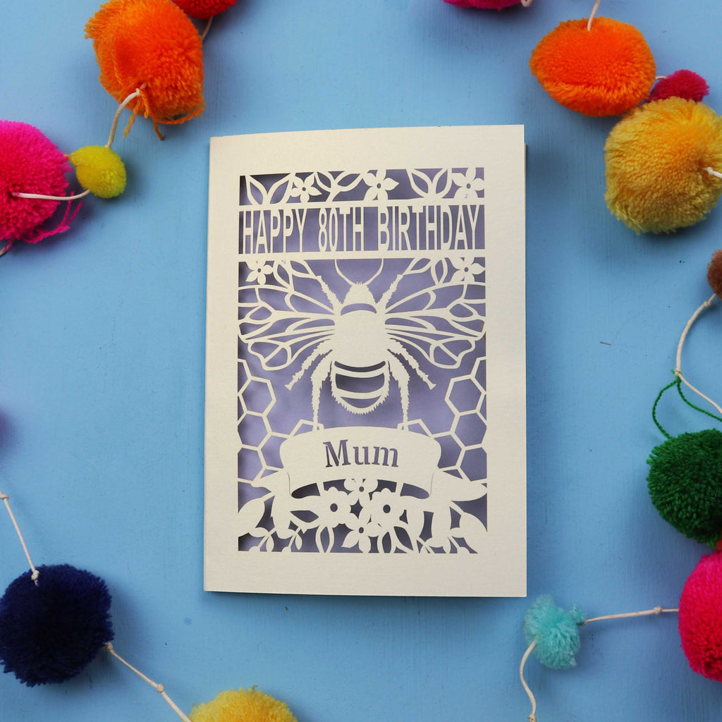 Laser-cut birthday card with bee design and 'Happy 80th Birthday Mum' text on a blue background with colourful pom-poms.
