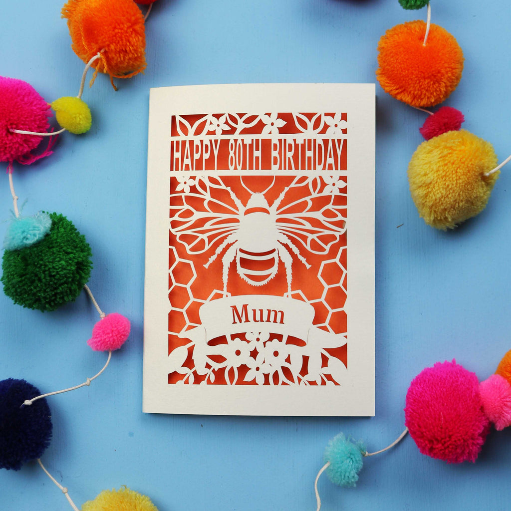 Laser-cut birthday card with bee design and 'Happy 80th Birthday Mum' text on a blue background with colourful pom-poms.