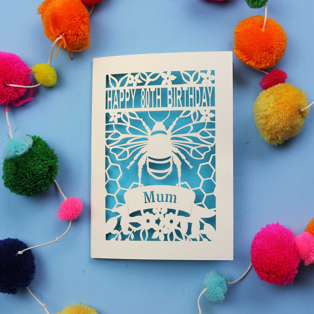 Laser-cut birthday card with bee design and 'Happy 80th Birthday Mum' text on a blue background with colourful pom-poms.