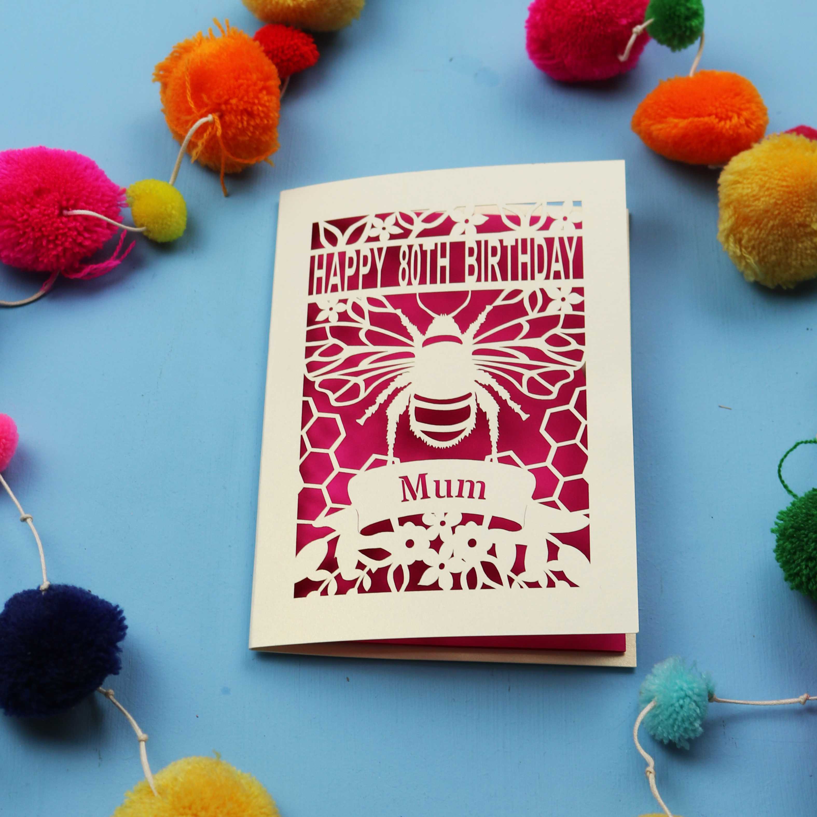 Laser-cut birthday card with bee design and 'Happy 80th Birthday Mum' text on a blue background with colourful pom-poms.