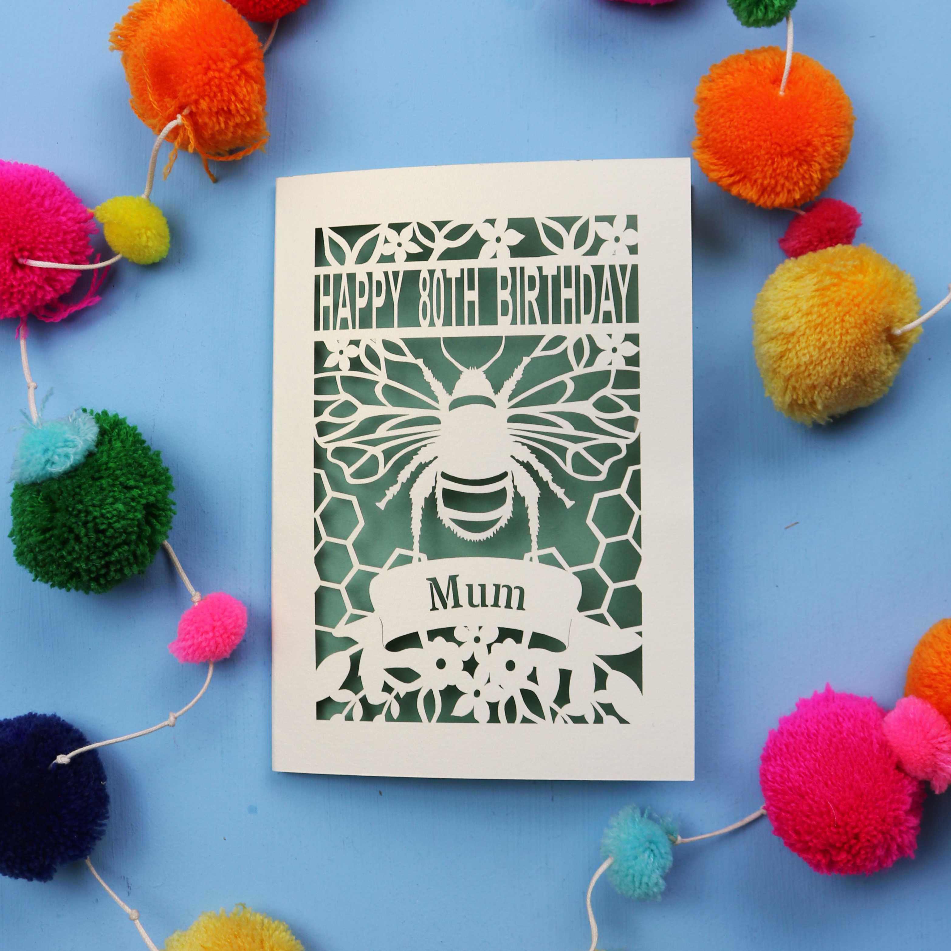 Laser-cut birthday card with bee design and 'Happy 80th Birthday Mum' text on a blue background with colourful pom-poms.