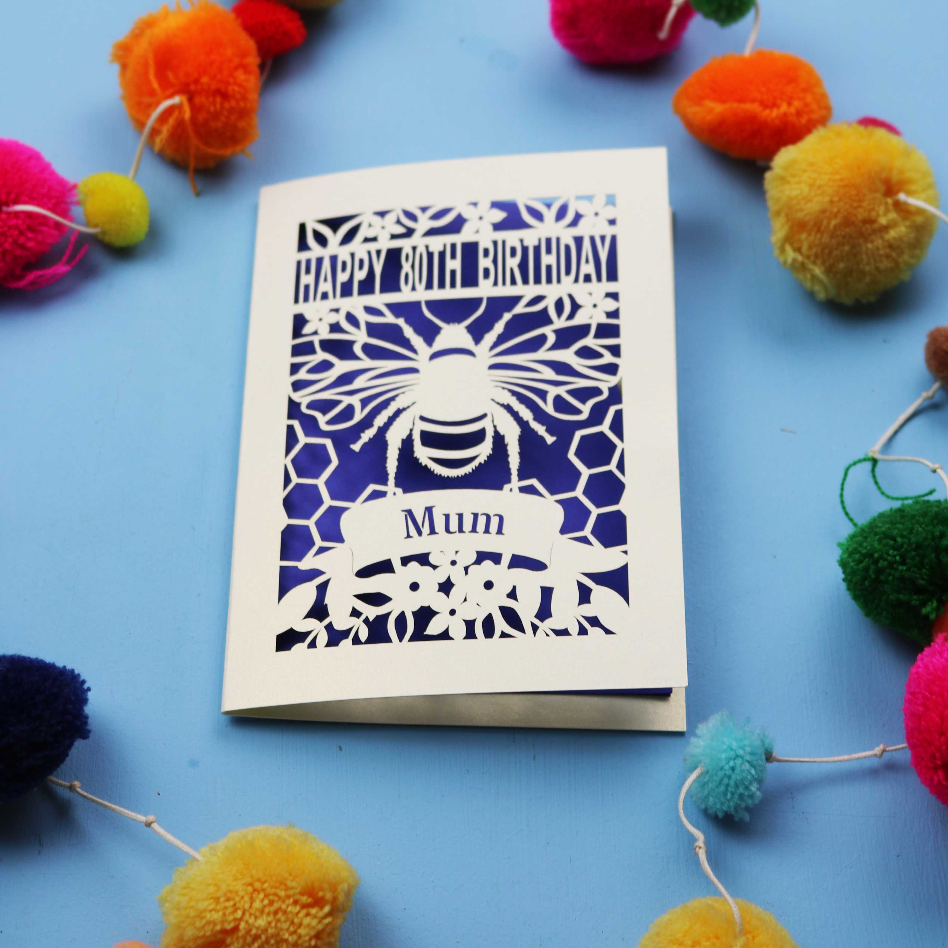 Laser-cut birthday card with bee design and 'Happy 80th Birthday Mum' text on a blue background with colourful pom-poms.