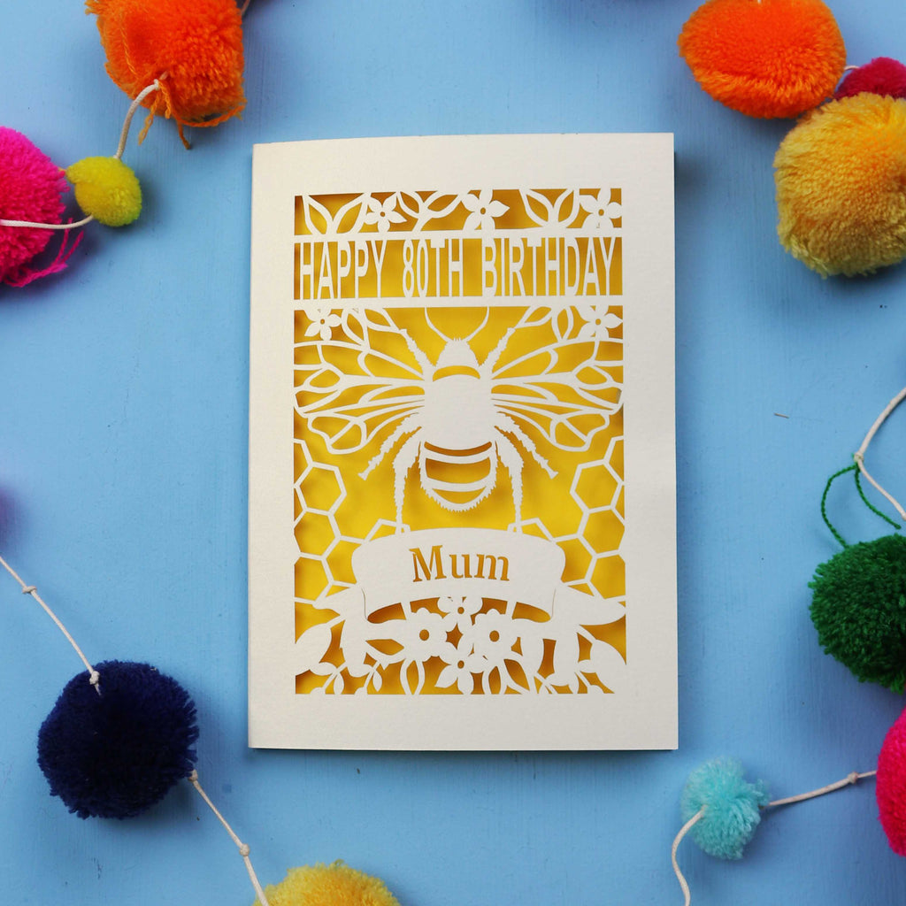 Laser-cut birthday card with bee design and 'Happy 80th Birthday Mum' text on a blue background with colourful pom-poms.