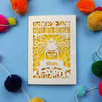 Laser-cut birthday card with bee design and 'Happy 80th Birthday Mum' text on a blue background with colourful pom-poms.