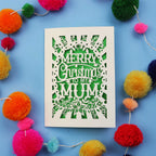 Papercut Christmas card with 'Merry Christmas to my Mum' text surrounded by colourful pom-poms on a blue background