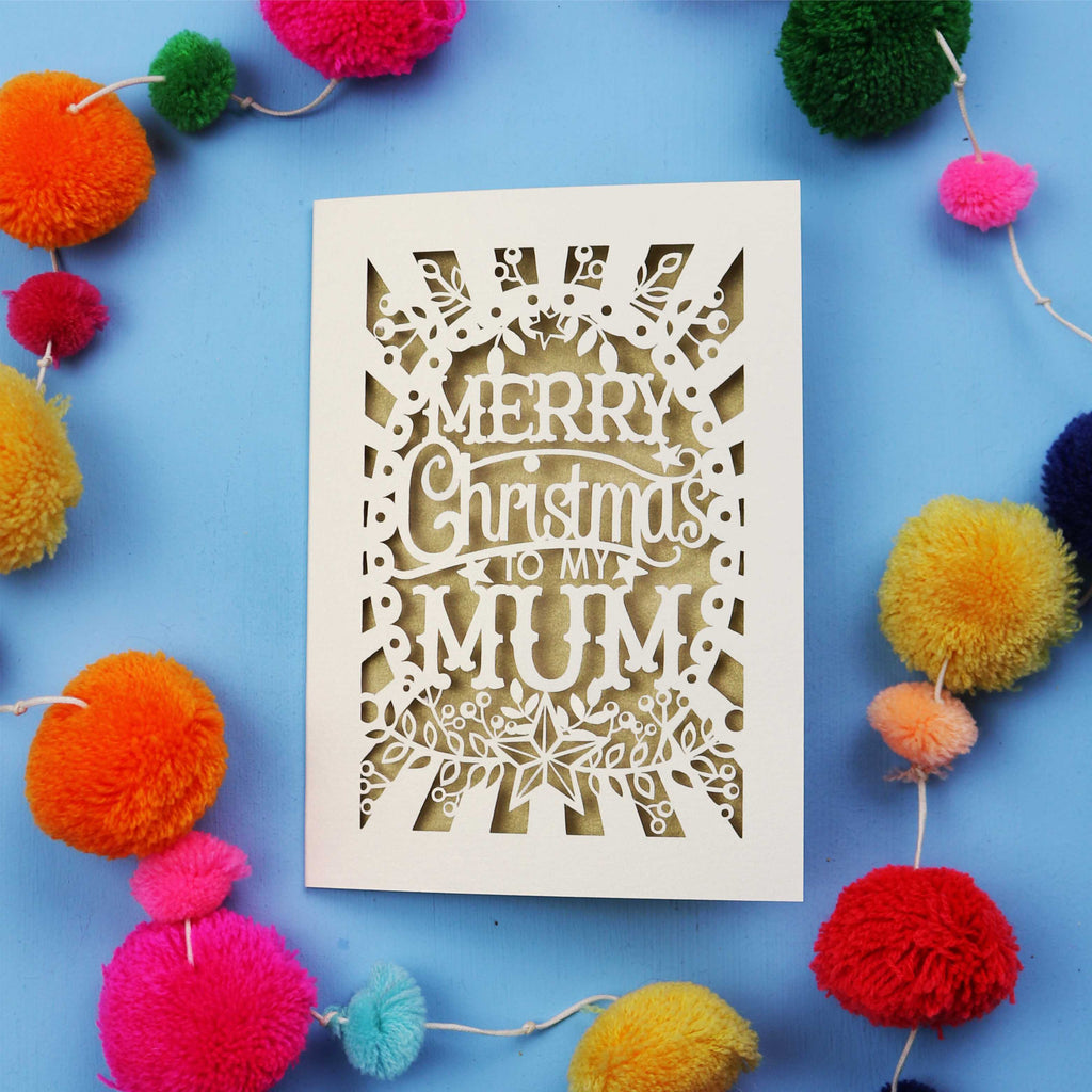 Decorative Laser Cut Christmas card with 'Merry Christmas to my Mum' text surrounded by colourful pom-poms on a blue background
