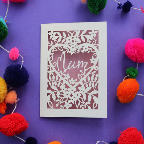A laser cut cream greetings card with flowers and leaves around a heart shape. The word "Mum" is inside the heart