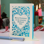 A cream personalised husband Christmas card with a laser-cut heart and floral pattern over a blue background, reading 'Merry Christmas to my amazing Husband Jake'. The card is displayed on a table with a blue pen in the foreground and blurred candles in the background.