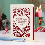 A cream personalised husband Christmas card with a laser-cut heart and floral pattern over a red background, reading 'Merry Christmas to my amazing Husband Jake'. The card is displayed on a table with a blue pen in the foreground and blurred candles in the background.