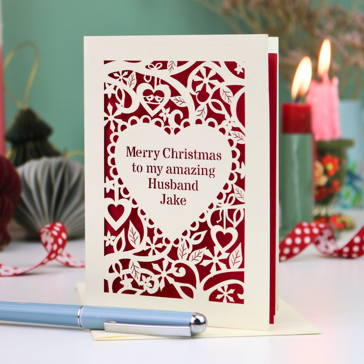 A cream personalised husband Christmas card with a laser-cut heart and floral pattern over a red background, reading 'Merry Christmas to my amazing Husband Jake'. The card is displayed on a table with a blue pen in the foreground and blurred candles in the background.