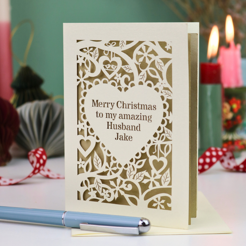A cream personalised husband Christmas card with a laser-cut heart and floral pattern over a gold background, reading 'Merry Christmas to my amazing Husband Jake'. The card is displayed on a table with a blue pen in the foreground and blurred candles in the background.