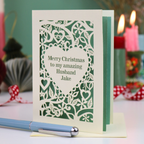 A cream personalised husband Christmas card with a laser-cut heart and floral pattern over a sage green background, reading 'Merry Christmas to my amazing Husband Jake'. The card is displayed on a table with a blue pen in the foreground and blurred candles in the background.