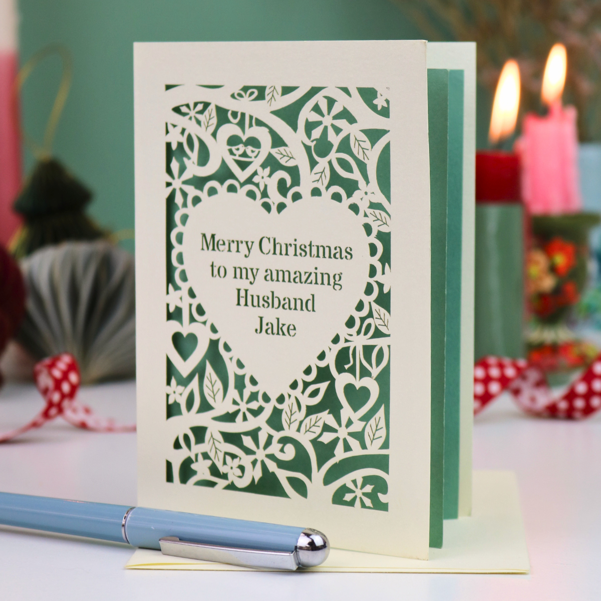 A cream personalised husband Christmas card with a laser-cut heart and floral pattern over a sage green background, reading 'Merry Christmas to my amazing Husband Jake'. The card is displayed on a table with a blue pen in the foreground and blurred candles in the background.