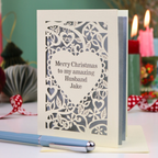 A cream personalised husband Christmas card with a laser-cut heart and floral pattern over a silver background, reading 'Merry Christmas to my amazing Husband Jake'. The card is displayed on a table with a blue pen in the foreground and blurred candles in the background.