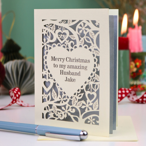 A cream personalised husband Christmas card with a laser-cut heart and floral pattern over a silver background, reading 'Merry Christmas to my amazing Husband Jake'. The card is displayed on a table with a blue pen in the foreground and blurred candles in the background.