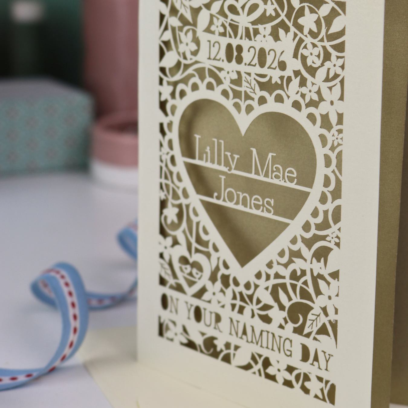 A laser cut 'On your naming day' card, personalised with a name and date, standing on a desk with stationery items.