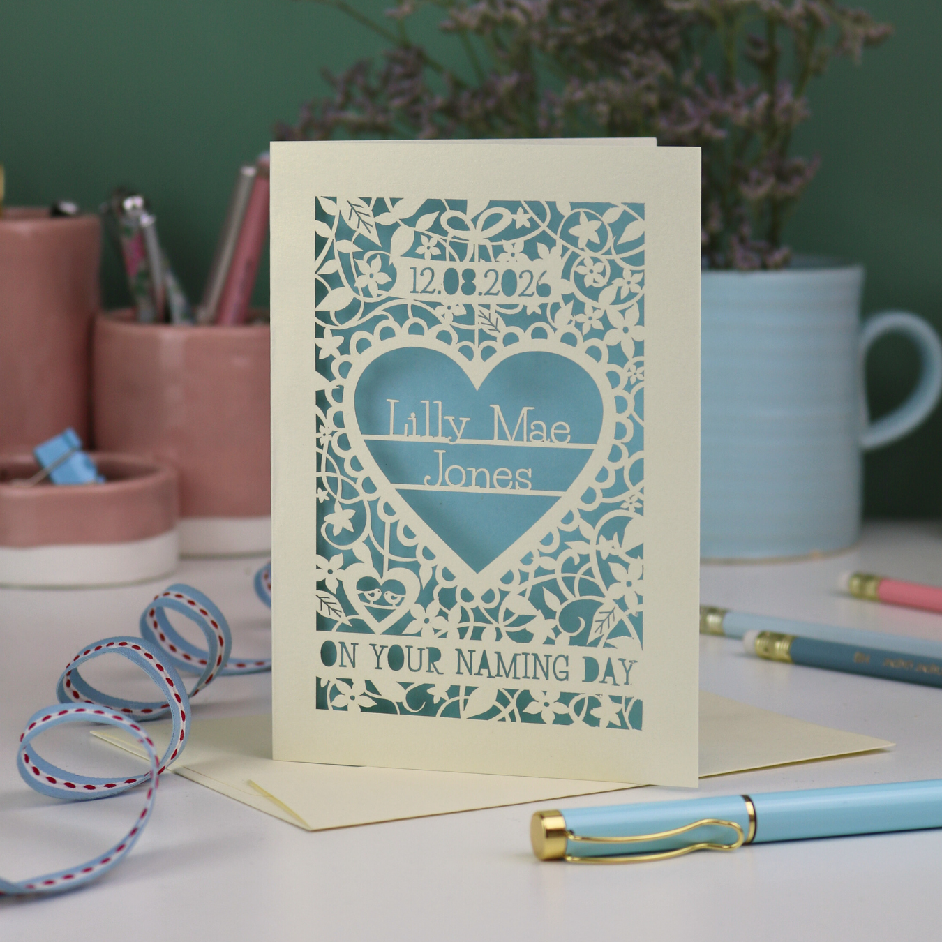 A keepsake laser cut 'On your naming day' card, personalised with a name and date, standing on a desk with stationery items.