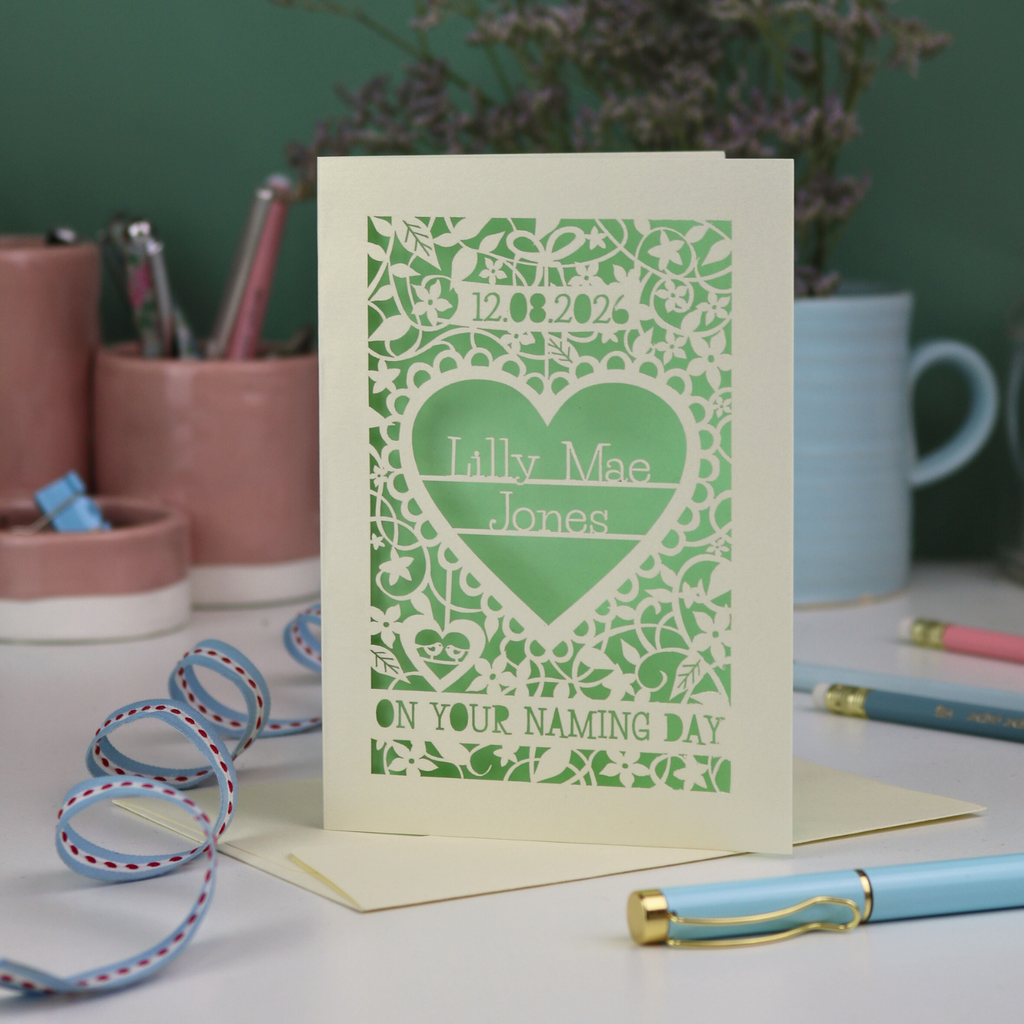 A unique laser cut 'On your naming day' card, personalised with a name and date, standing on a desk with stationery items.