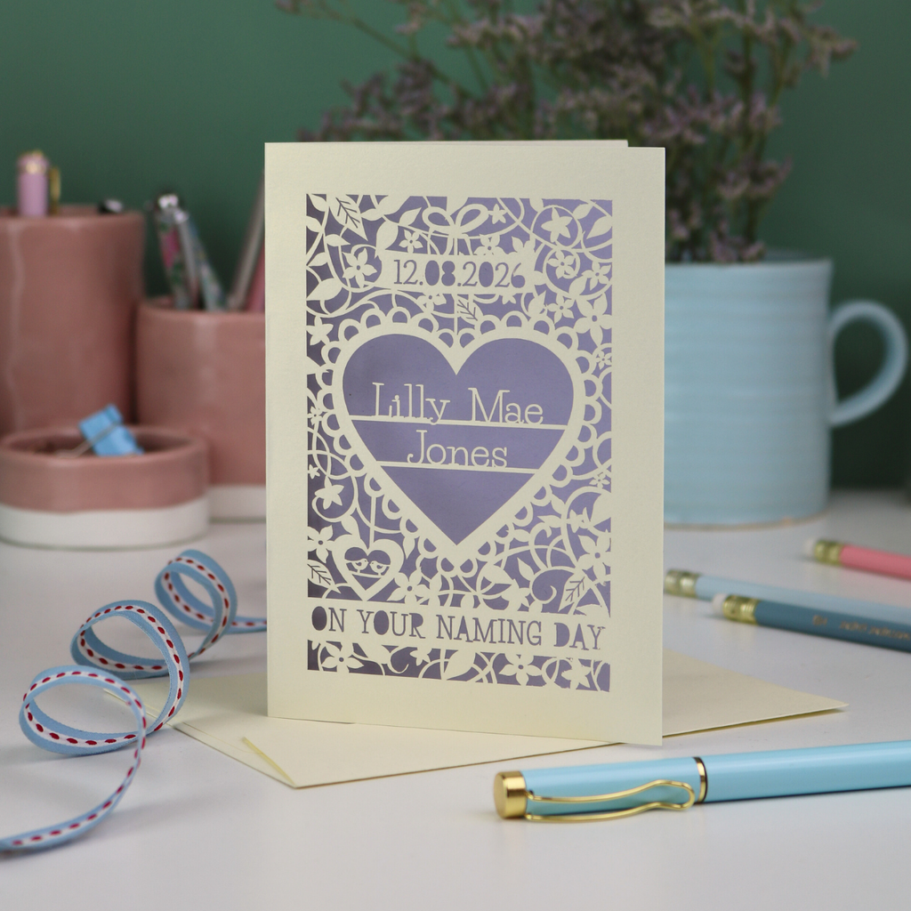 A laser cut 'On your naming day' card, personalised with a name and date, standing on a desk with stationery items.