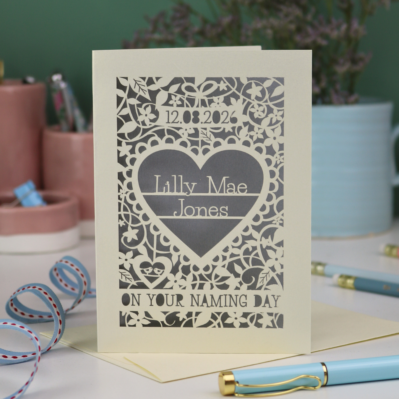 A laser cut 'On your naming day' card, personalised with a name and date, standing on a desk with stationery items.