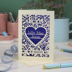A laser cut 'On your naming day' card, personalised with a name and date, standing on a desk with stationery items.