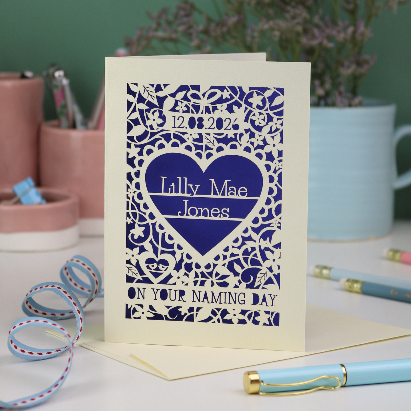 A laser cut 'On your naming day' card, personalised with a name and date, standing on a desk with stationery items.