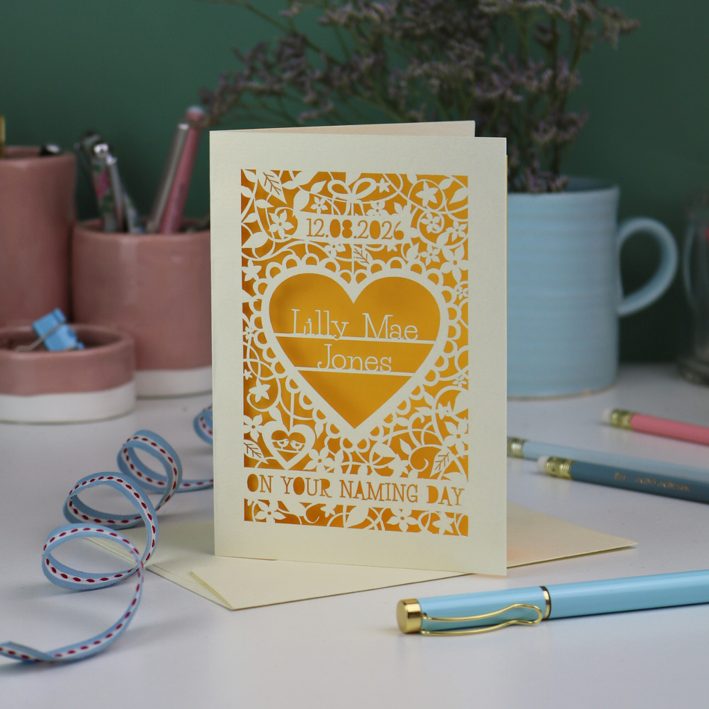 A laser cut and personalised 'On your naming day' card, personalised with a name and date, standing on a desk with stationery items.