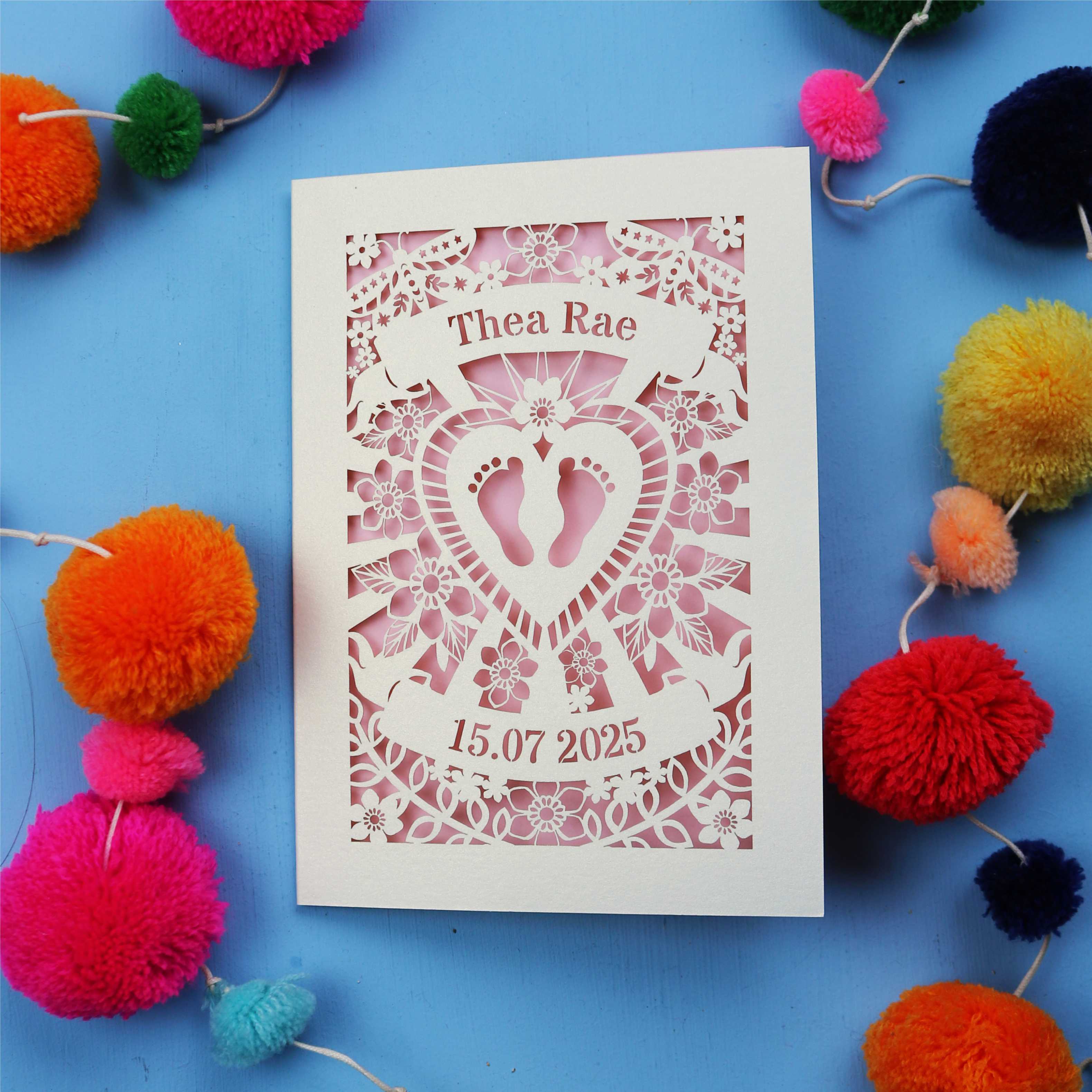 Laser-cut baby card with personalised text on a blue background with colourful pom-poms.