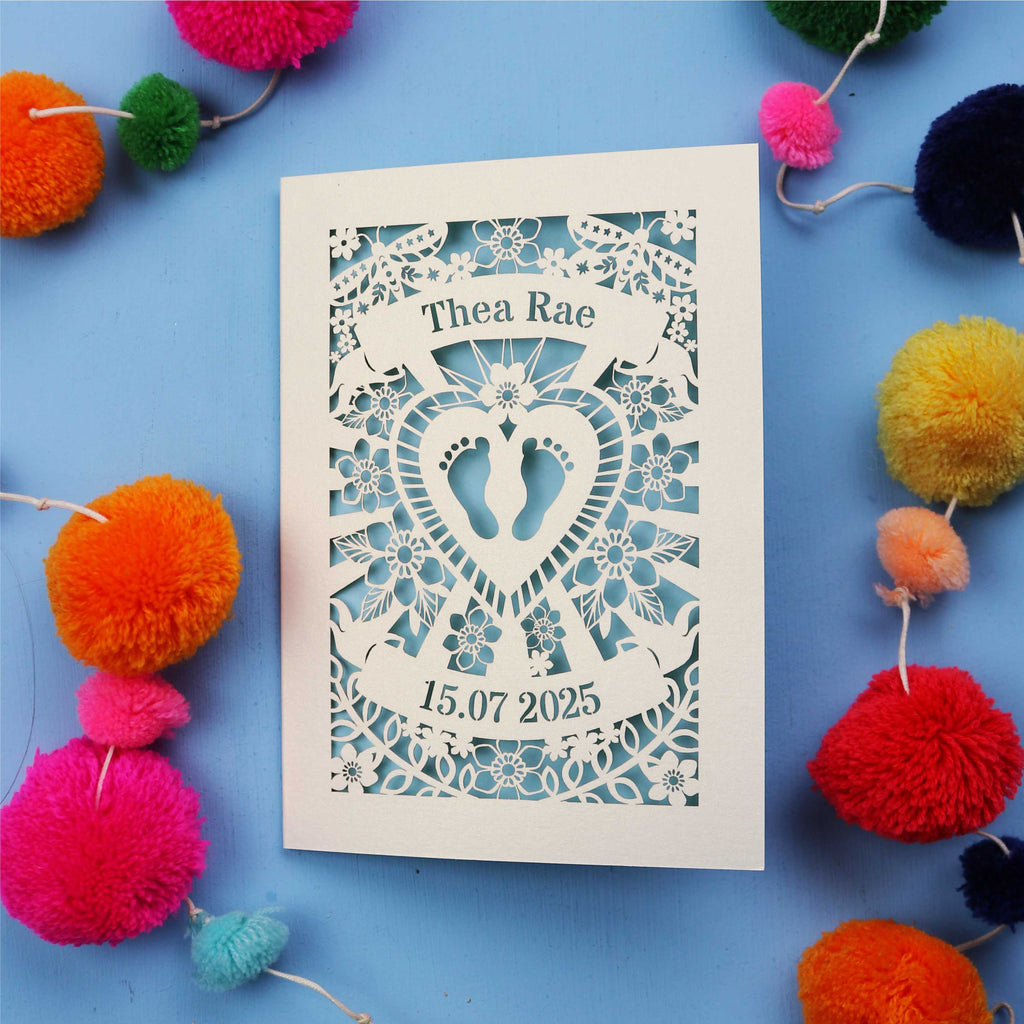 Personalised baby card with personalised name and date, and intricate laser cut design on a blue background with colourful pom-poms.