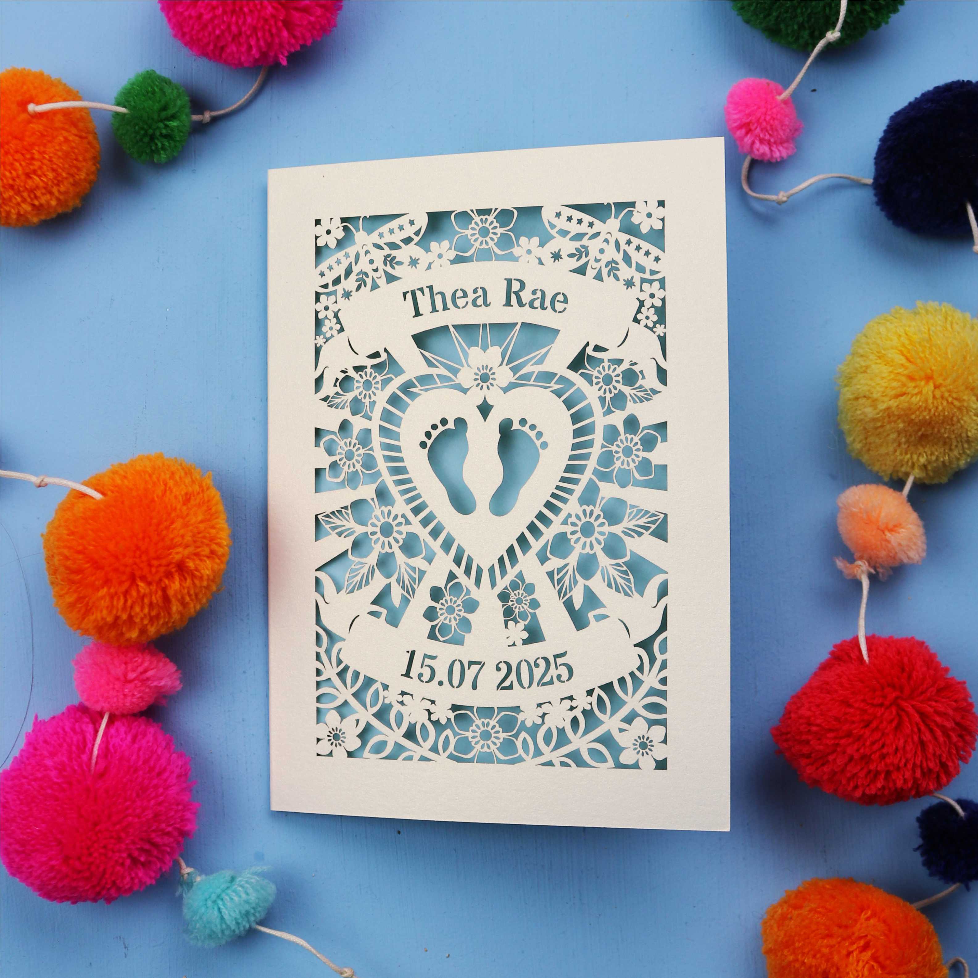 Personalised baby card with personalised name and date, and intricate laser cut design on a blue background with colourful pom-poms.