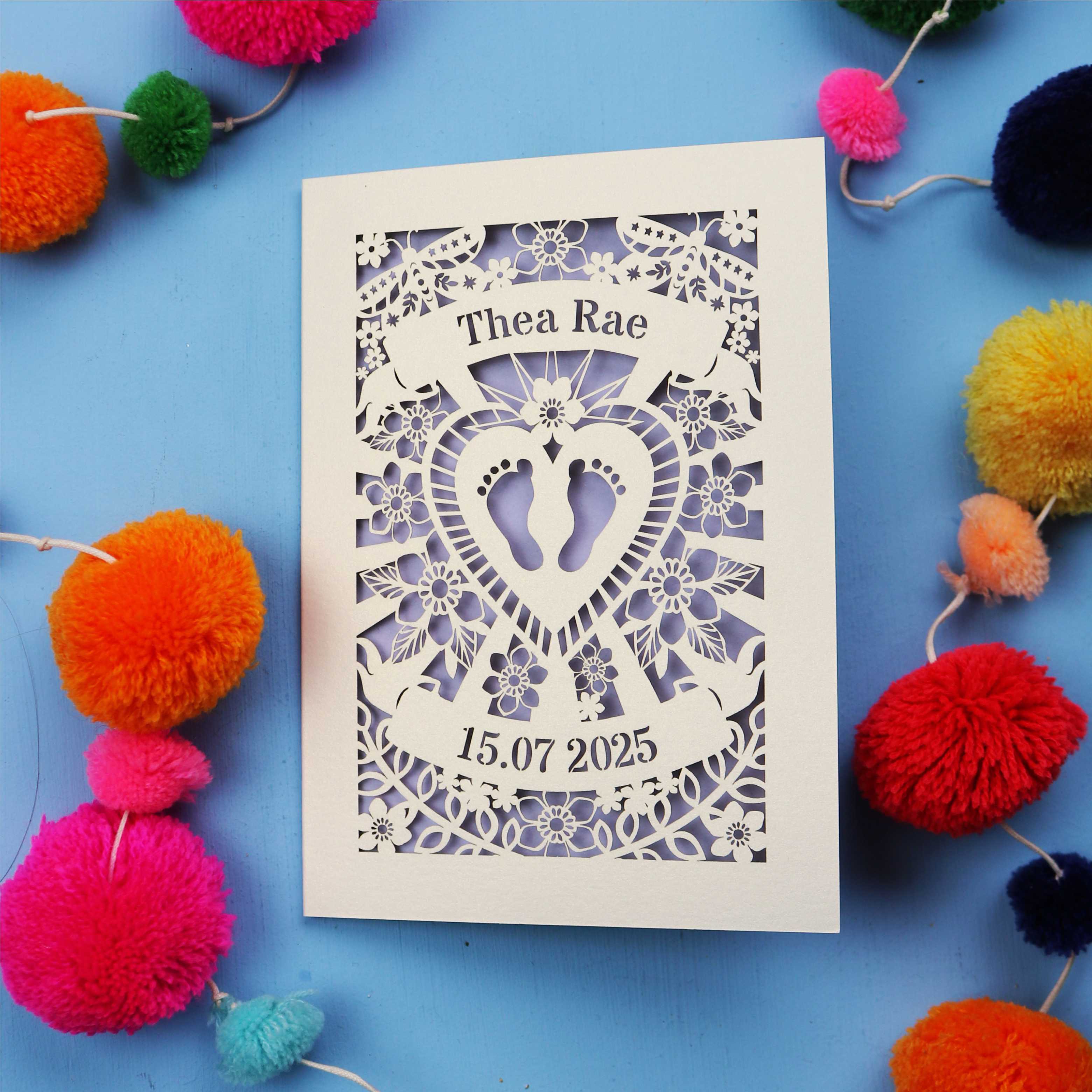Personalised cut out baby card with personalised name and date, and intricate laser cut design on a blue background with colourful pom-poms.