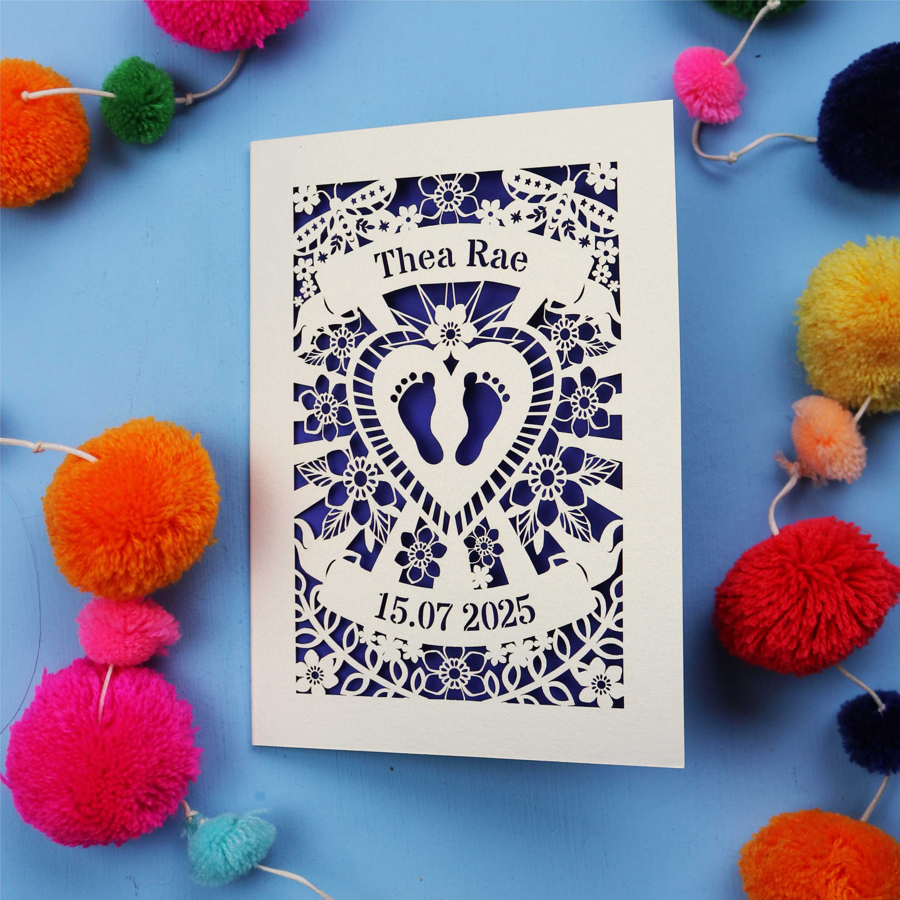 Keepsake Personalised baby card with personalised name and date, and intricate laser cut design on a blue background with colourful pom-poms.