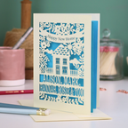 Laser-cut housewarming card with personalized names on a table with stationery items.