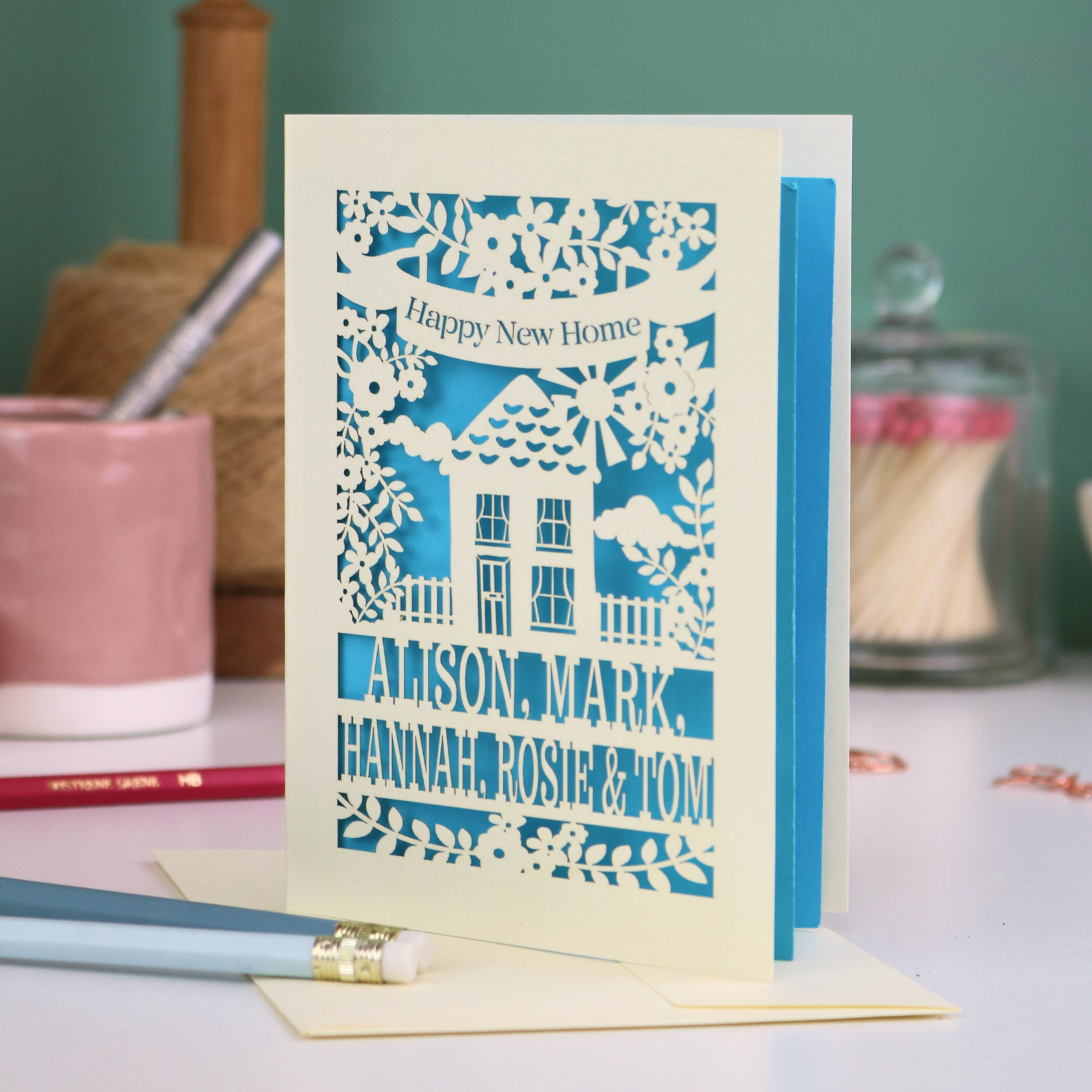 Laser-cut housewarming card with personalized names on a table with stationery items.