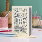 Laser-cut 'Happy New Home' card with a house design on a table with stationery items.