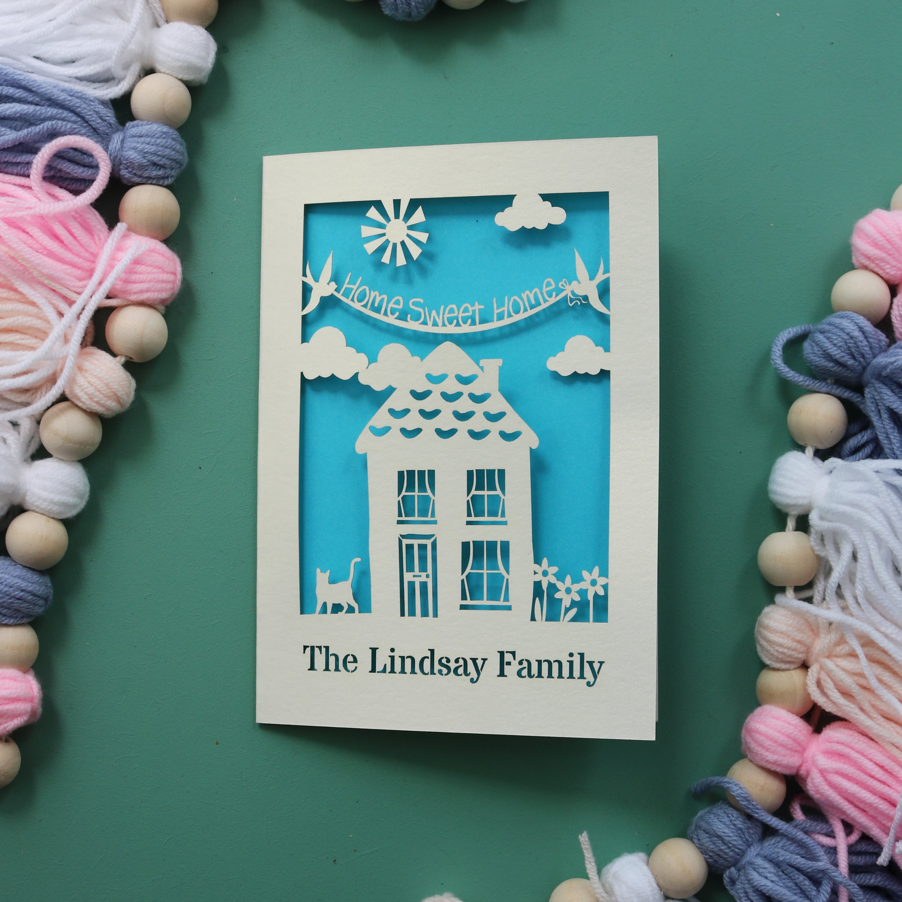 A cream and blue paper cut housewarming card with a house, cat and flowers, and the words "Home sweet home". Card is personalised at the bottom with a line of text.