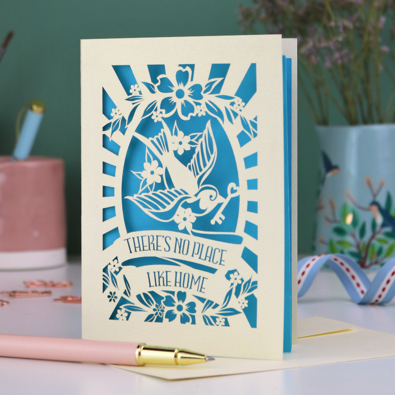 A laser cut new home card with cut out swallow, key and flower design and text in a banner, "There's no place like home"