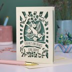 A laser cut new home card with cut out swallow design and text in a banner, "There's no place like home"
