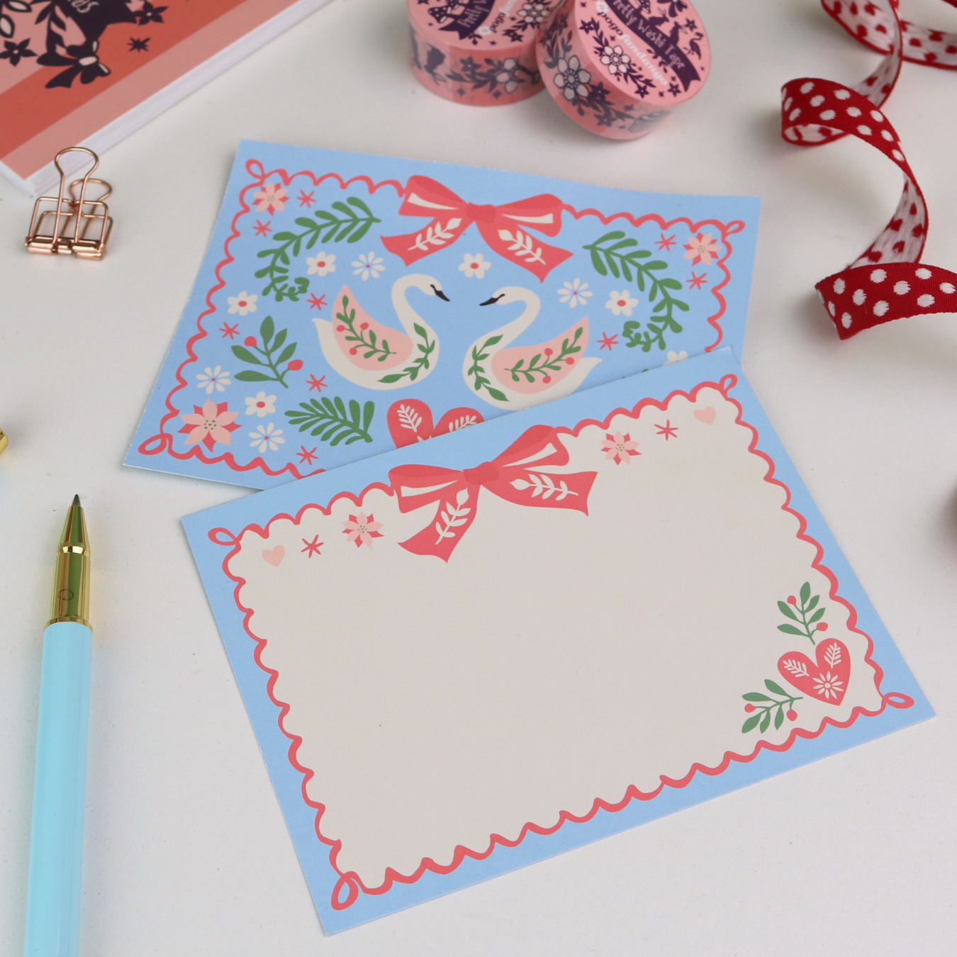 Two decorative note cards with swan designs on a white surface with ribbons and a pen.