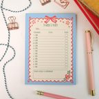 A flat-lay view of a 'Today's Plan' notepad with a light blue background and pink/red border details, styled on a white surface with a pink pen, bobby pin clips, and string.