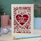 A cream laser cut engagement card on a desk with stationery items. The card is portrait-orientation, with floral cut out details and 'Congratulations, name and name, on your engagement'