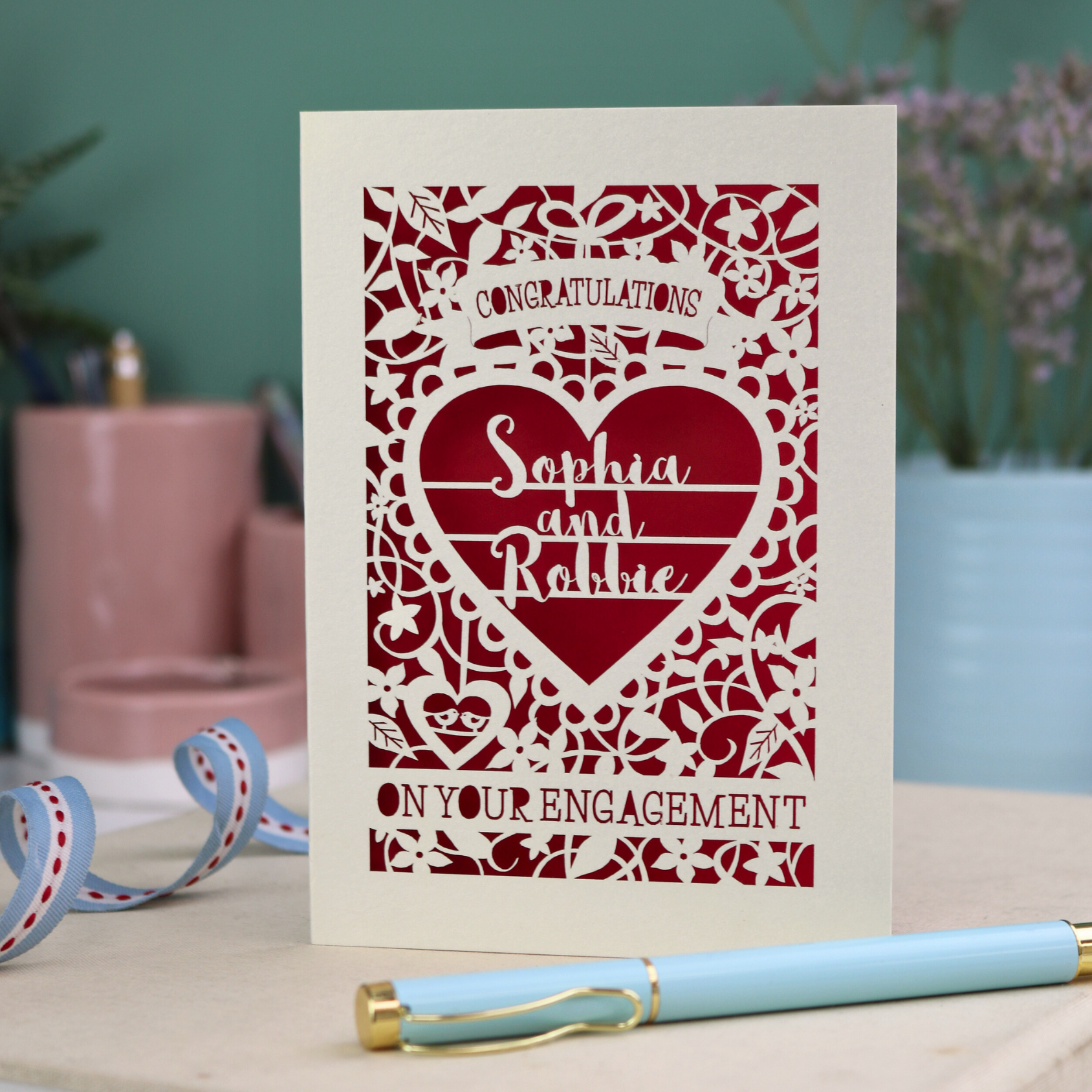 A cream laser cut engagement card on a desk with stationery items. The card is portrait-orientation, with floral cut out details and 'Congratulations, name and name, on your engagement'