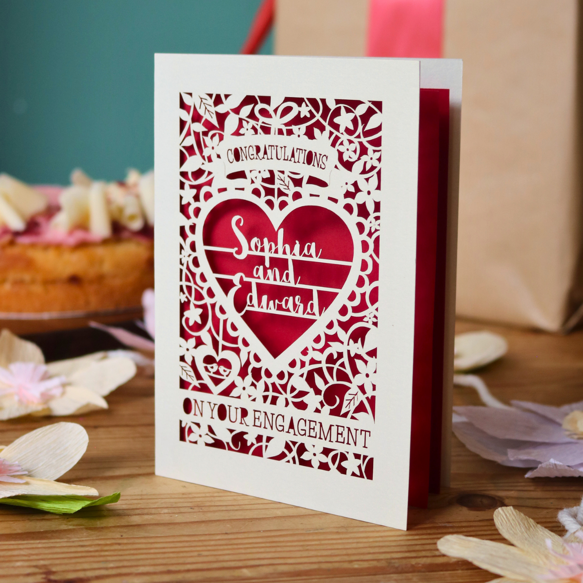 A cream laser cut engagement card with intricate floral border and heart design. Inside the heart are the names 'Sophia and Edward' in a brush script font, above is the word 'Congratulations', and below 'on your engagement'