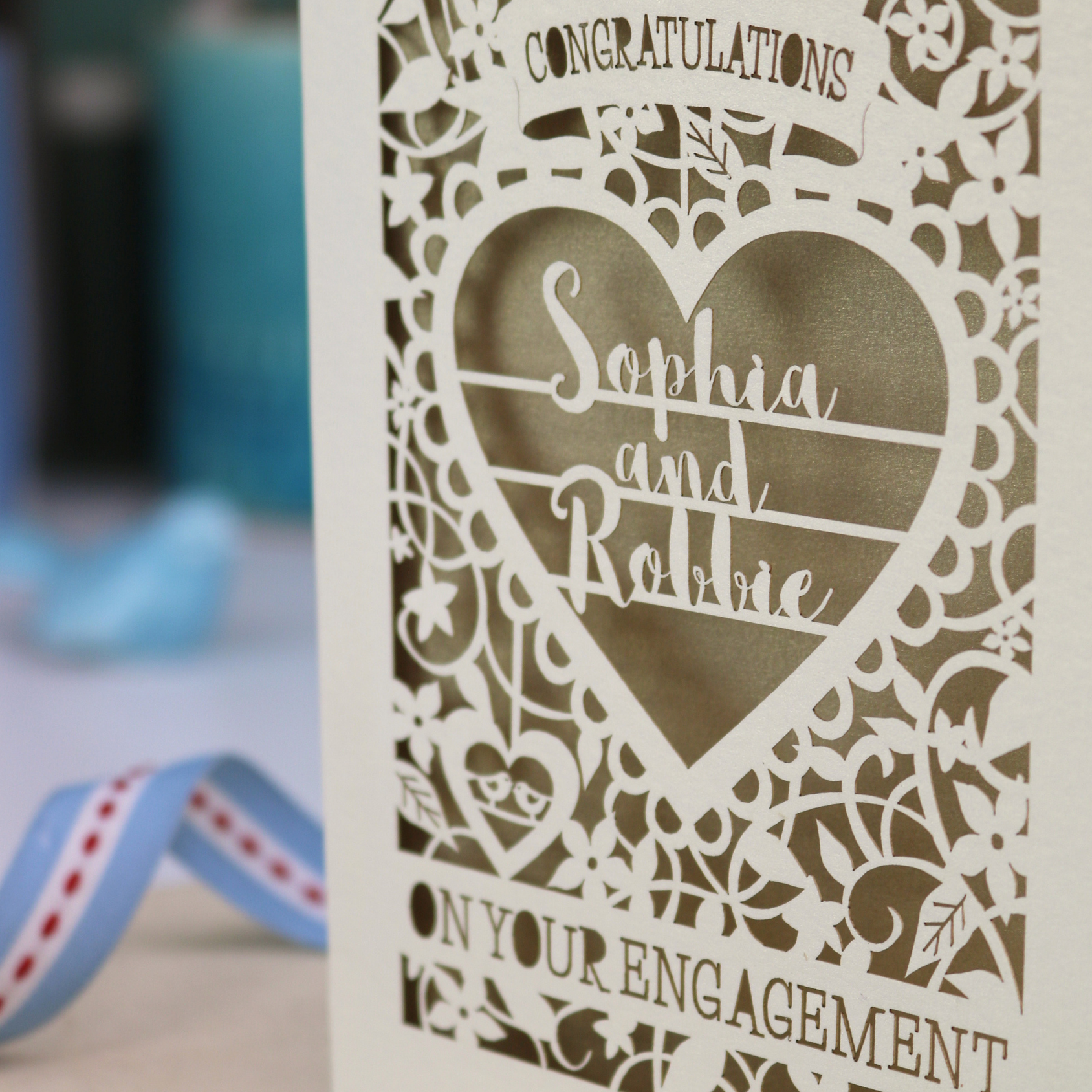 A cream laser cut engagement card on a desk with stationery items. The card is portrait-orientation, with floral cut out details and 'Congratulations, name and name, on your engagement'