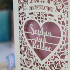 A cream laser cut engagement card on a desk with stationery items. The card is portrait-orientation, with floral cut out details and 'Congratulations, name and name, on your engagement'
