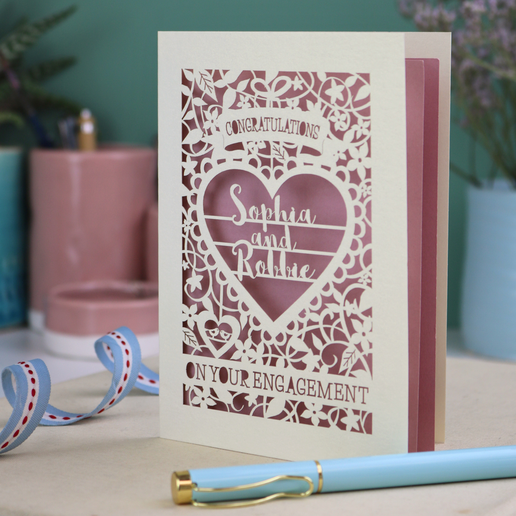 A cream laser cut engagement card on a desk with stationery items. The card is portrait-orientation, with floral cut out details and 'Congratulations, name and name, on your engagement'