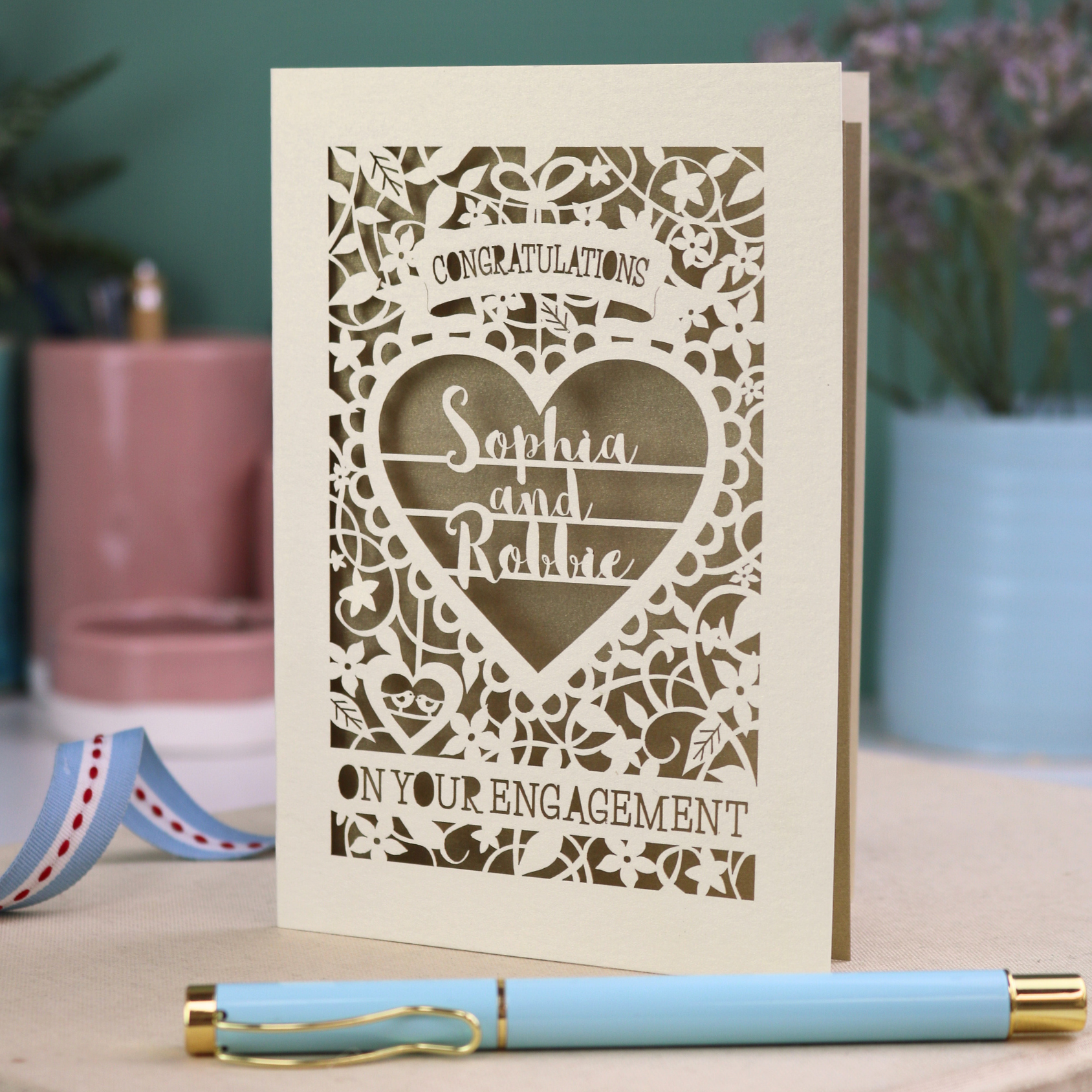 A cream laser cut engagement card on a desk with stationery items. The card is portrait-orientation, with floral cut out details and 'Congratulations, name and name, on your engagement'